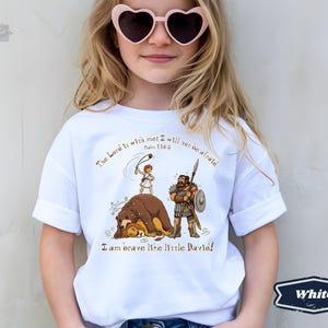 Op de afbeelding: Wit T-shirt met een cartoon illustratie van David en Goliath. Het shirt heeft de tekst "De Heer is met mij; ik zal niet bang zijn" en "Ik ben dapper als kleine David!" in een decoratief lettertype.