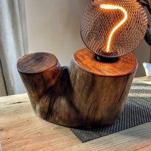 Puede incluir: Una lámpara de madera única con un acabado marrón oscuro. La lámpara presenta un globo de malla metálica con el número siete brillante en su interior. La lámpara se asienta sobre una base de madera natural, añadiendo un toque rústico al diseño.