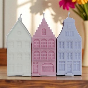 Könnte beinhalten: Drei dekorative Kerzen in Hausform in Weiß, Rosa und Lavendel. Jede Kerze hat ein anderes architektonisches Design mit Fenstern und Türen. Im Hintergrund ist eine Vase mit Blumen zu sehen.