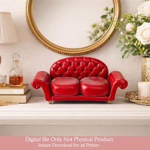 May include: A miniature red sofa with a button-tufted back and two seat cushions. The sofa is a decorative item, possibly for a dollhouse or display. The background includes a gold-framed mirror, a lamp, and a vase of white roses.