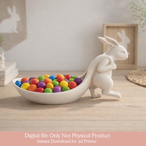 Könnte beinhalten: Eine weiße Keramik-Hasenfigur, die eine weiße ovale Schale mit bunten Süßigkeiten trägt. Der Hase steht auf seinen Hinterbeinen, wobei sich die Schale von seinem Rücken erstreckt. Die Süßigkeiten sind in verschiedenen Farben, darunter Rot, Gelb, Blau und Lila.