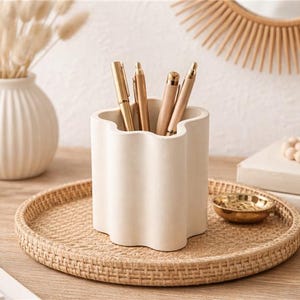 May include: A neutral-toned desk arrangement featuring a cream-colored pen holder with gold pens, a woven tray, a small gold bowl, and a decorative vase with dried flowers. A wooden sunburst mirror is in the background.