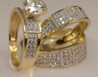Könnte beinhalten: Drei goldfarbene Ringe, zwei davon mit einem Band aus kleinen, funkelnden Steinen. Der größte Ring hat einen prominenten, klaren, oval geschliffenen Stein. Die Ringe sind vor einem weichen, neutralen Hintergrund platziert, der ihr Design hervorhebt.