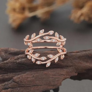 May include: Rose gold ring featuring a double band design. Each band is adorned with delicate leaf-shaped accents, embellished with small, sparkling gemstones. The ring is set against a dark, textured background, highlighting its intricate details.
