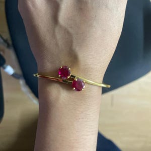 May include: A gold-toned bypass bangle bracelet featuring two round, faceted, deep pinkish-red stones set in prong settings at the open ends of the band, creating a symmetrical design that rests across the wrist.