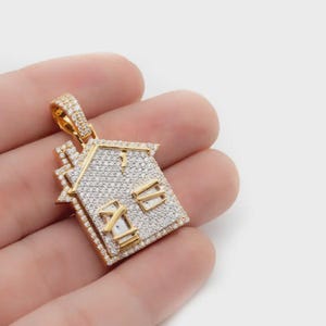 Puede incluir: Un colgante dorado con forma de casa, incrustado con pequeñas piedras blancas brillantes. El colgante tiene un diseño detallado de techo y ventanas. La argolla también está cubierta con las mismas piedras blancas. El colgante se sostiene en una mano.