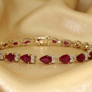 May include: A gold bracelet with pear-shaped, deep red gemstones and small white accent stones. The bracelet is set against a gold-colored, draped fabric, creating a luxurious feel. The clasp is visible in the center.