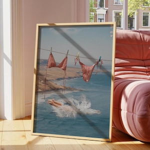 May include: Framed print showing a red and white checkered bikini hanging on a washing line above a coastal scene. A person dives into the sea, creating a splash. The artwork is displayed in a room with a pink sofa and wooden floor.