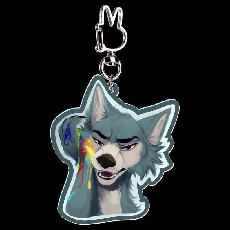 May include: A keychain featuring a stylized wolf character with blue fur and a stern expression. The wolf has colorful paint streaks on its face. The keychain has a silver-colored clasp and a bunny-shaped charm.