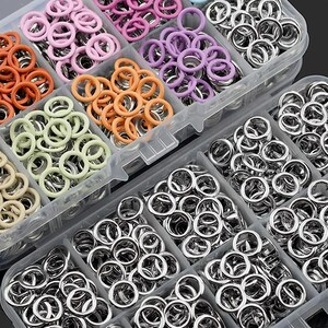May include: A clear plastic organizer filled with various sizes and colors of metal eyelets. The eyelets have colored rims in shades of orange, pink, purple, and green. The majority of the eyelets are silver.