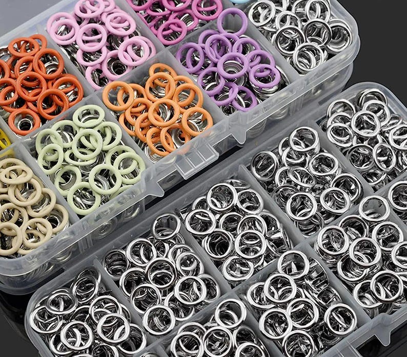 May include: A clear plastic organizer filled with various sizes and colors of metal eyelets. The eyelets have colored rims in shades of orange, pink, purple, and green. The majority of the eyelets are silver.
