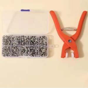 May include: A set of silver-colored metal snap fasteners organized in a clear plastic storage container with ten compartments, accompanied by a pair of orange handheld pliers featuring a metal spring mechanism and white rubberized tips.