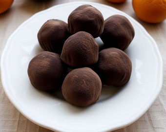 May include: A white plate holds a cluster of seven dark chocolate truffles. The truffles are round, dusted with cocoa powder, and have a slightly rough texture. Oranges and a wooden bowl with orange segments are in the background.