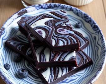 May include: Dark chocolate bars with white and blue swirls are arranged on a blue and white patterned plate. Fresh blueberries are scattered around the plate and in a small white bowl. A bottle with a blueberry label is in the background.