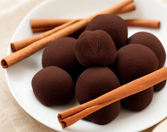 May include: A white plate holds several dark brown, spherical treats, possibly truffles or candies, alongside several cinnamon sticks. The treats have a textured surface. The cinnamon sticks are a light brown color.