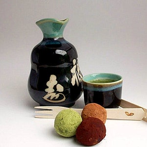 May include: A Japanese tea set with a dark blue ceramic sake bottle and cup, both with teal accents. The cup contains green tea. Three truffles and a wooden fork and tongs complete the arrangement.