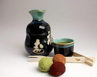 May include: A Japanese tea set with a dark blue ceramic sake bottle and cup, both with teal accents. The cup contains green tea. Three truffles and a wooden fork and tongs complete the arrangement.