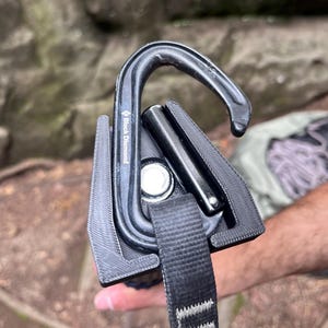 May include: Black Diamond climbing carabiner with a black strap. The carabiner is black and has the Black Diamond logo. The strap is black and has white stitching. The carabiner is designed for climbing and mountaineering.