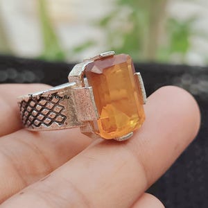 Pode incluir: Um anel de prata com uma grande pedra preciosa retangular de cor âmbar. O anel tem uma banda texturizada e as palavras "Sai Jewelry Crafts" são visíveis na lateral. A pedra preciosa é engastada em uma moldura de prata.