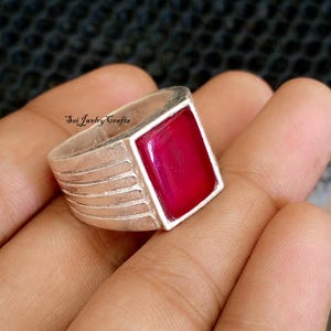 Peut inclure: Une bague en argent avec une pierre rouge foncé rectangulaire. La bague a une bande épaisse avec des lignes horizontales sur le côté. La bague est tenue dans une main. Le texte "Sai Jewelry Crafts" est visible.