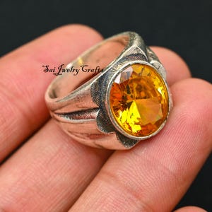 May include: A silver ring with an oval, yellow gemstone. The ring has a textured band and a decorative setting around the stone. The text "Sai Jewelry Crafts" is visible on the ring.