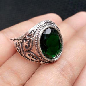 May include: Silver-toned ring with an oval, emerald-green gemstone. The ring features an ornate, engraved design around the band and the gemstone setting. The text "Sai Jewelry Crafts" is visible on the side of the ring.