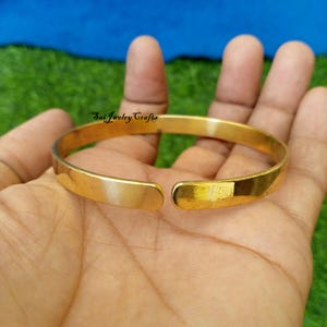 May include: A gold-colored, open cuff bracelet. The bracelet is made of a solid metal and has a smooth, polished finish. The text "Sai Jewelry Crafts" is visible on the bracelet. The bracelet is shown on a hand.