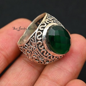 Pode incluir: Um anel de prata com uma pedra preciosa verde esmeralda oval e facetada. O anel tem um padrão ornamental e ondulado ao redor da banda. O texto "Sai Jewelry Crafts" é visível.