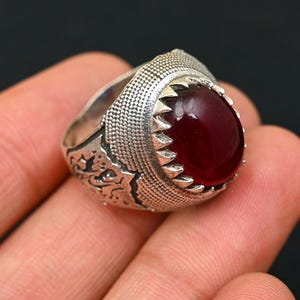 May include: A silver ring with an oval, deep red gemstone. The ring has an ornate, textured band with a detailed pattern. The gemstone is set in a serrated bezel, adding to the ring's vintage aesthetic.