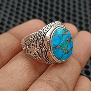 Può includere: Anello in argento con pietra turchese ovale. La pietra turchese è di un blu brillante con venature marroni e dorate. L'anello presenta un design ornato e dettagliato sulla fascia.