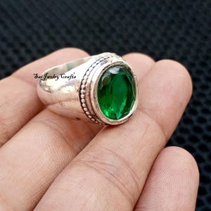 以下が含まれることがあります： 楕円形のエメラルドグリーンの宝石が付いたシルバーリング。リングにはビーズのデザインと「Sai Jewelry Crafts」の文字が入った詳細なバンドが付いています。宝石はベゼルセッティングでセットされており、リングは手の上に表示されています。