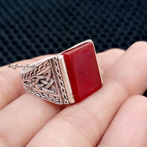May include: A silver ring with a rectangular, deep red stone. The ring's band features an intricate Celtic knot design. The ring is a close-up shot, showcasing the details of the metalwork and the stone's colour.