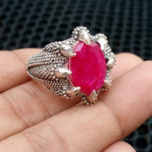 May include: A silver ring featuring a large, oval-cut, vibrant red gemstone. The ring has a textured, dotted band and claw-like prongs securing the stone. The design is intricate and detailed, showcasing a unique, statement piece of jewelry.