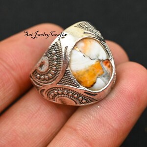 May include: A silver-toned ring with an oval-shaped, white, orange, and gray stone. The ring has an ornate, textured design around the stone and on the band. The text "Sai Jewelry Crafts" is visible on the ring.