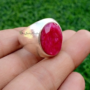 May include: A silver ring with a large, oval-cut ruby gemstone. The ruby is a vibrant red color and is set in a polished silver band. The ring has the text "Sai Jewelry Crafts" engraved on the side.