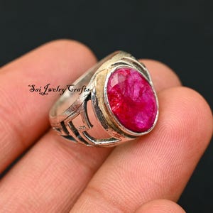 Op de afbeelding: Zilveren ring met een grote, ovaal geslepen robijn. De ring heeft een decoratief, open ontwerp aan de zijkanten en een goudkleurige band rond de robijn. De tekst "Sai Jewelry Crafts" is zichtbaar.