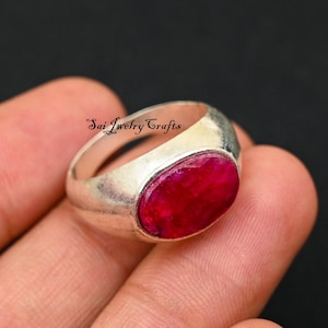 May include: A silver ring with a large, oval-shaped, deep red gemstone. The ring has a wide band and a polished finish. The text "Sai Jewelry Crafts" is visible on the ring. The ring is shown on a finger.