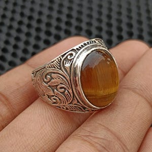 Op de afbeelding: Een zilveren ring met een ovale tijgeroogsteen. De ring heeft een sierlijk, gegraveerd ontwerp op de band. De tijgeroogsteen heeft een warme bruine kleur met gouden strepen.