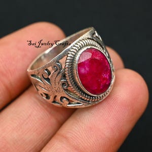 Puede incluir: Anillo de plata con una gema ovalada de color rojo intenso. El anillo tiene un diseño ornamental de filigrana en la banda y alrededor de la gema. El texto "Sai Jwelry Crafts" es visible en el anillo.