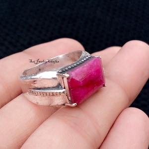 May include: A silver ring with a large, rectangular, faceted, deep pink gemstone. The ring has a textured band and the text "Sai Jewelry Crafts" is visible on the side.