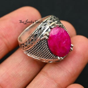 Puede incluir: Un anillo de plata con una gema rosa brillante de forma ovalada. El anillo tiene intrincados diseños de nudos celtas en la banda y el texto "Sai Jewelry Crafts". El anillo se sostiene en una mano.