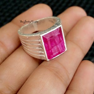 May include: A silver ring with a large, rectangular, magenta-colored gemstone. The ring band has a ribbed design. The text "Sai Jewelry Crafts" is visible on the ring.