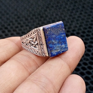 Può includere: Un anello d'argento con una pietra rettangolare di lapislazzuli blu scuro. L'anello presenta un'intricata lavorazione a nodi celtici sulla fascia e il testo "Sai Jewel Crafts". La pietra ha macchie dorate.