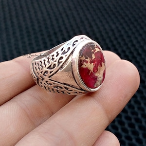 May include: Silver-toned ring with an oval, red stone featuring gold-colored inclusions. The ring has an ornate, openwork design on the band. The ring is held in a hand.