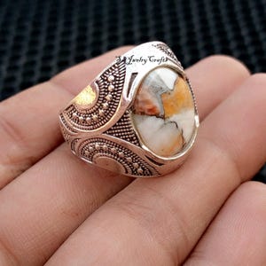 May include: Silver-toned ring with an oval, multi-colored stone. The stone features white, orange, and brown hues. The ring has an ornate design with dotted and curved patterns. The text "SJ Jewelry Crafts" is visible on the ring.