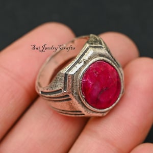 May include: A silver ring with a large, oval, deep red gemstone. The ring has a geometric design around the gemstone and the text "Sai Jewelry Crafts" is visible on the ring.