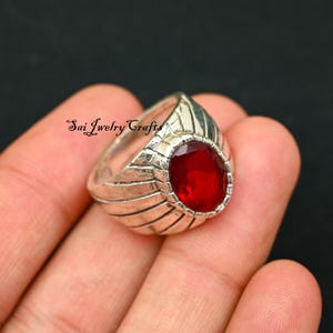 Peut inclure: Une bague en argent ornée d'une grande pierre rouge ovale. La bague présente un motif texturé sur le corps et le texte "Sai Jewelry Crafts". La pierre rouge est le point central, sertie dans un sertissage argenté détaillé.