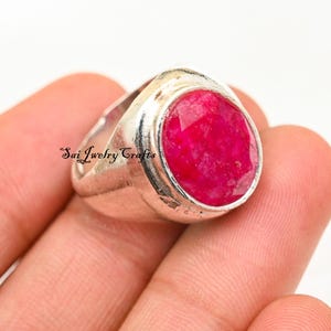 Può includere: Un anello d'argento con una grande pietra preziosa rossa brillante di forma ovale. L'anello ha una fascia spessa e un'incastonatura a castone. Il testo "Sai Jewelry Crafts" è stampato sull'anello.