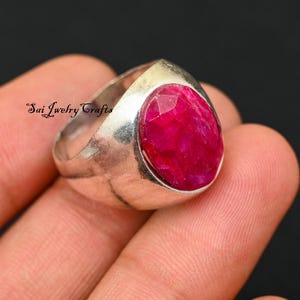 May include: A silver ring with a large, oval-cut ruby gemstone. The ruby is a vibrant red color and is set in a polished silver band. The ring is a statement piece of jewelry.