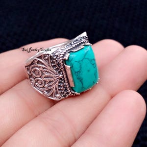 Puede incluir: Un anillo de plata con un diseño ornamentado y una gran piedra turquesa rectangular. El anillo presenta patrones detallados en la banda y alrededor de la piedra, creando una estética vintage o antigua. La piedra turquesa es de un color azul-verdoso vibrante.